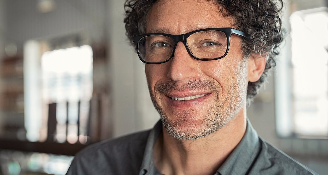 A man with curly hair and glasses smiles at the camera, wearing a gray shirt. The background is softly blurred.