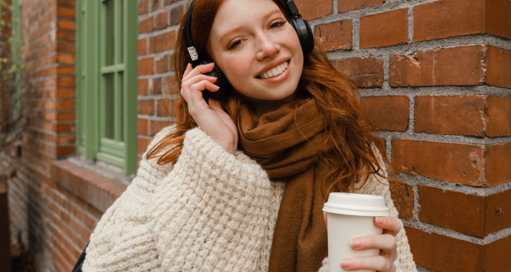 A person with red hair is wearing headphones and a scarf while holding a coffee cup, standing outside near a brick wall.