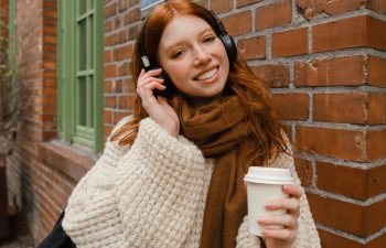 A person with red hair is wearing headphones and a scarf while holding a coffee cup, standing outside near a brick wall.